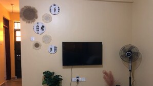 A living room with a flat-screen TV mounted on the wall, a standing fan, a potted plant, and decorative wall hangings.