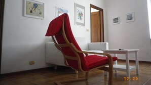 Una habitación con una mecedora de madera, un sofá blanco y una mesita. Hay cuadros enmarcados en las paredes.