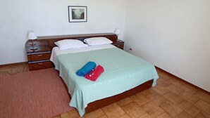 Una cama bien hecha con colcha verde y dos toallas enrolladas, una mesita de noche de madera con una lámpara y un cuadro en la pared.