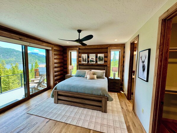 Un dormitorio con una cama grande, un ventilador de techo y vistas a un lago y montañas a través de amplios ventanales.
