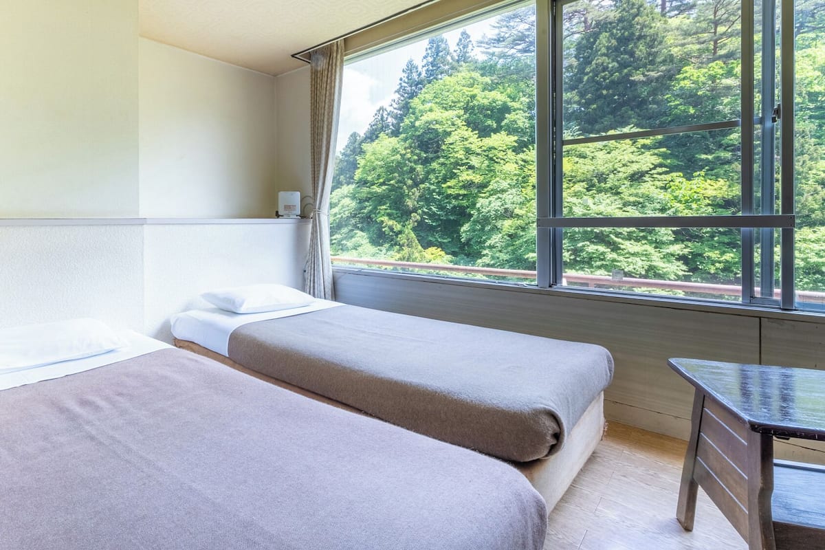 A room with two beds, a small table, and a window with a forest view.