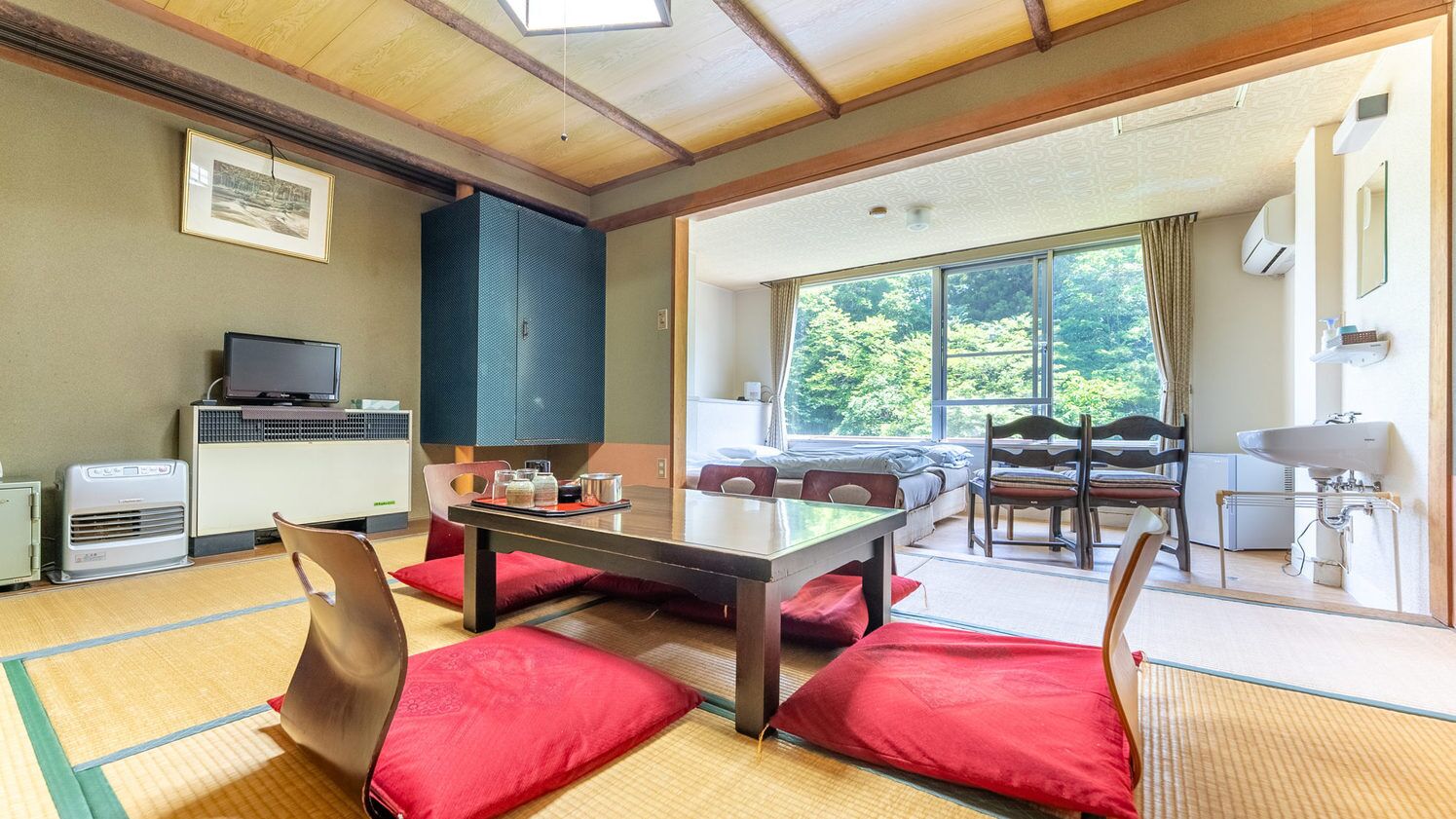 Een kamer met tatami matten, een lage tafel, stoelen, een televisie en een groot raam met uitzicht op bomen.