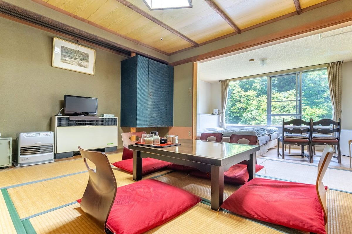 A room with tatami mats, a low table, chairs, a television, and a large window with a view of trees.