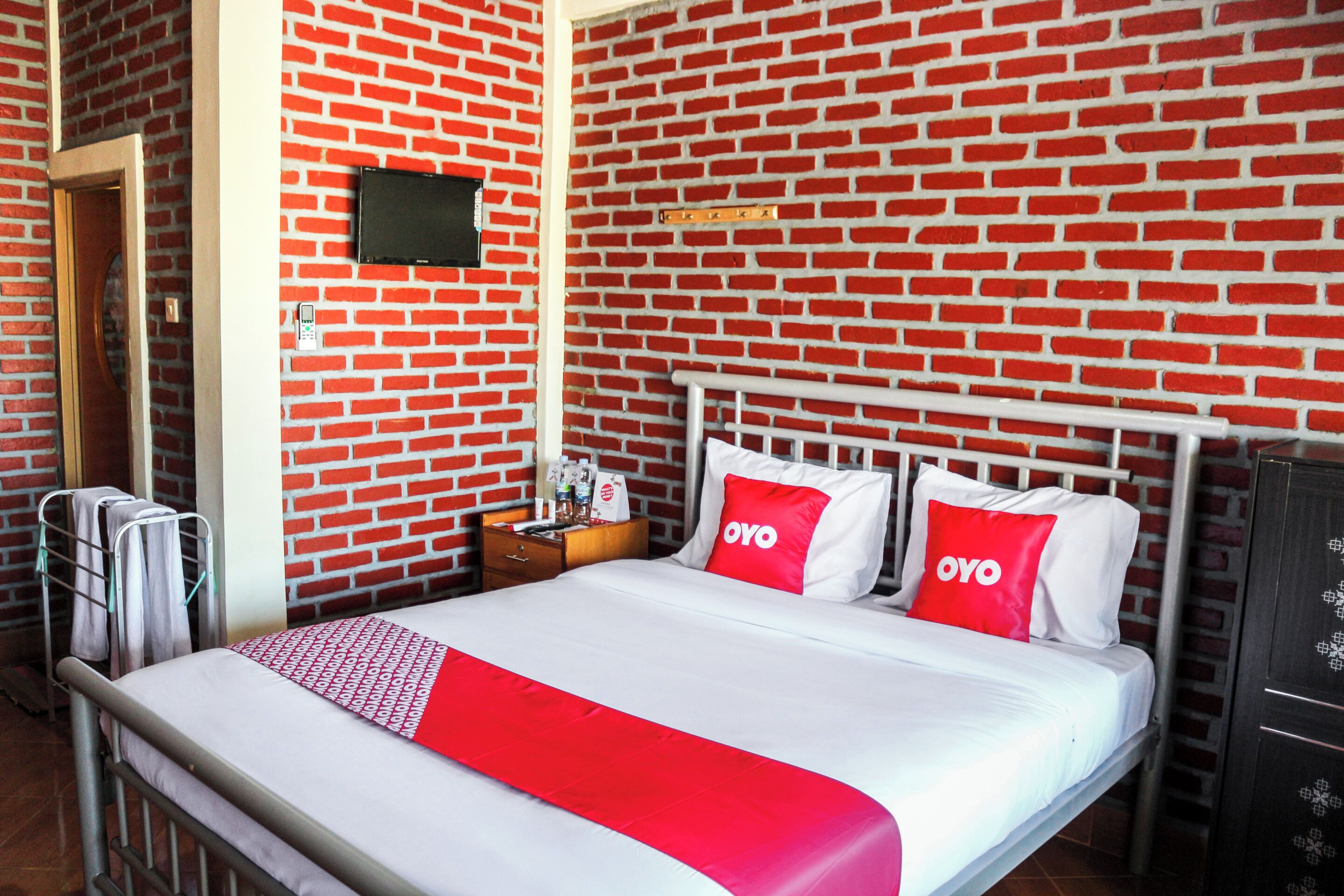 Habitación de hotel con pared de ladrillo, cama con ropa blanca y detalles rojos, televisor de pantalla plana sobre la cama y mesita de noche de madera con lámpara y una botella.