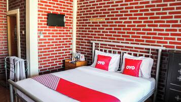 A hotel room with a brick-patterned wall, a bed with white bedding and red accents, a flat-screen TV mounted above the bed, and a wooden nightstand with a lamp and a bottle.