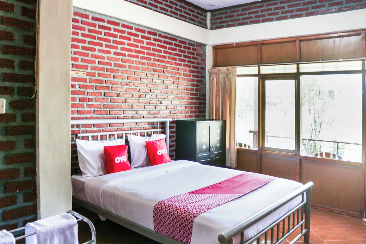 A single bed with a red and white patterned bedspread, a brick wall, a television on a stand, and a window with a view of the outside.