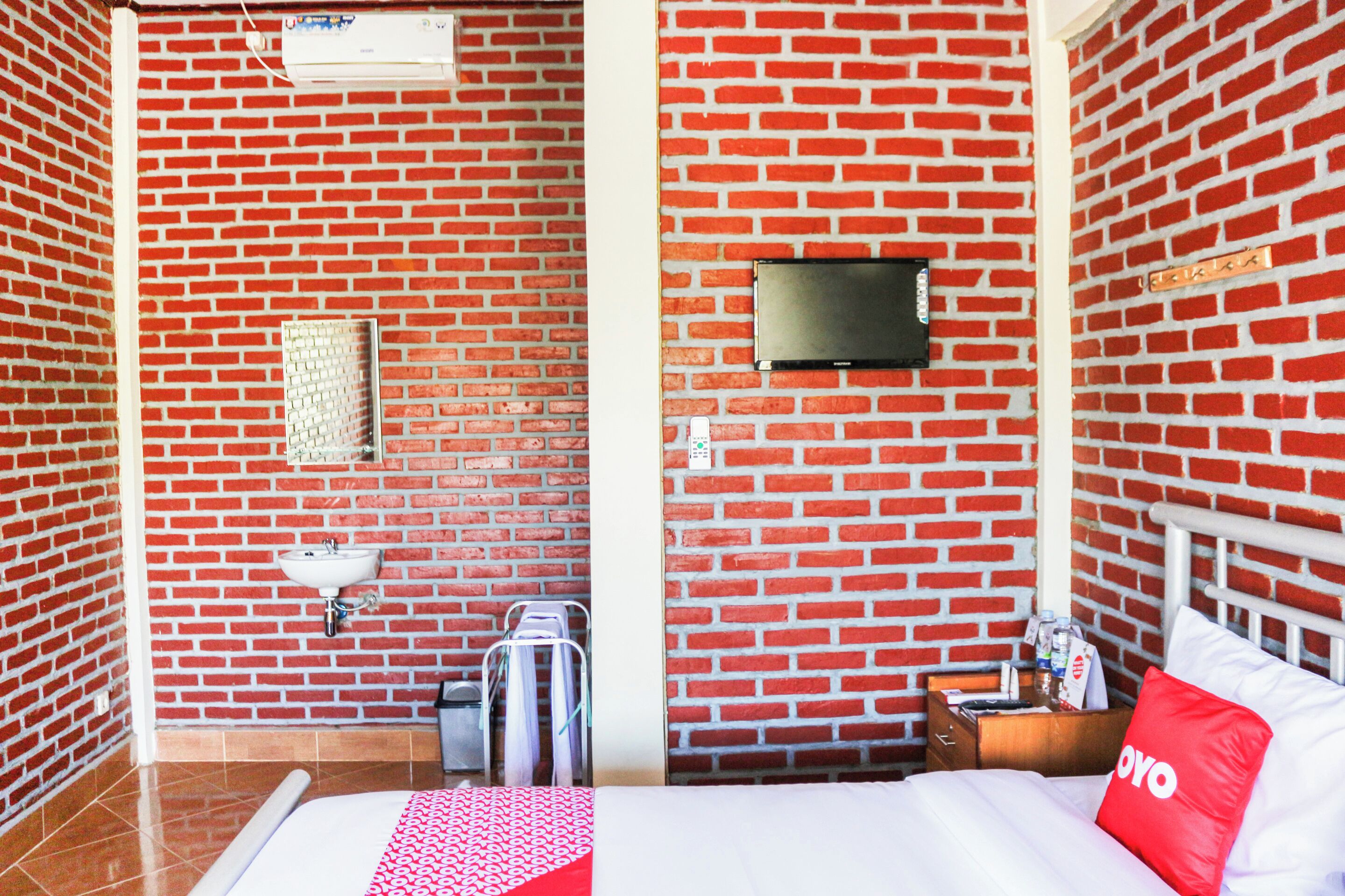 A hotel room with a brick wall, a TV mounted on the wall, a bed with a red pillow, a bedside table, and a small cabinet.