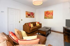 Un salón con un sofá marrón, una mesa de centro y un mueble para televisión. Hay dos cuadros en la pared, una puerta y una lámpara.