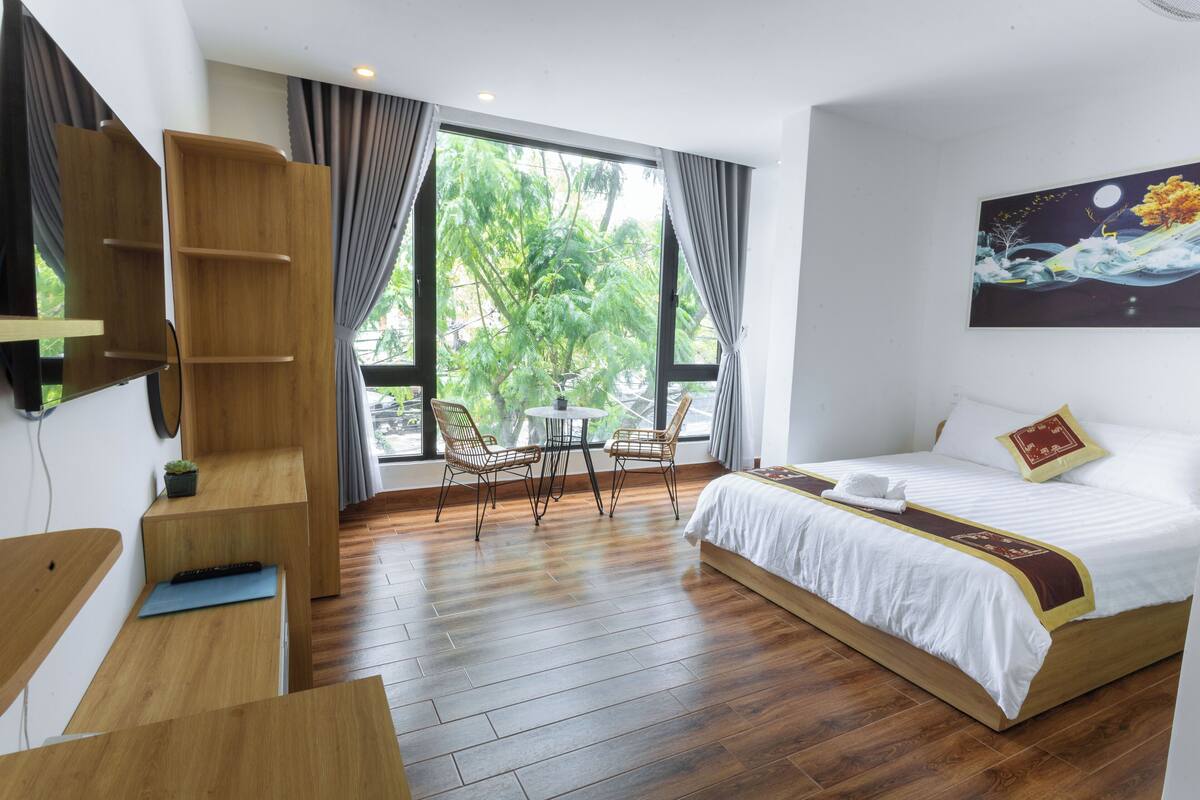A hotel room with a bed, a desk, a chair, and a window with a view of trees.