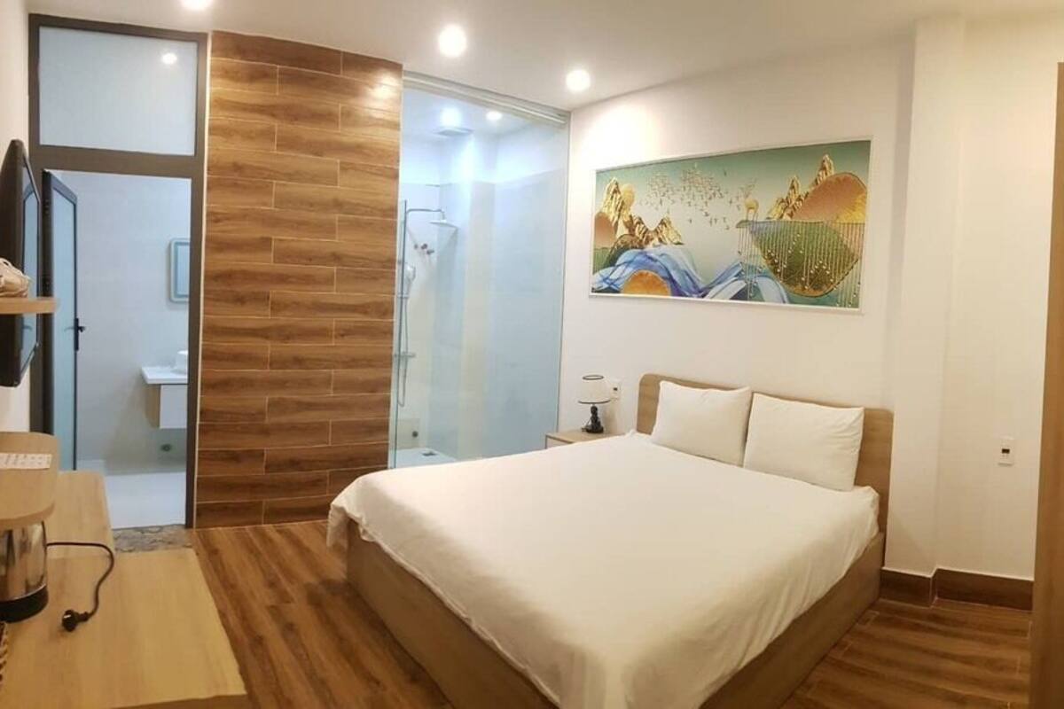 A hotel room with a bed, a wooden shelf with towels, a painting on the wall, and a glass shower area.