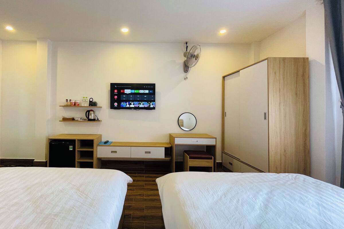 A hotel room with two beds, a desk with a chair, a wardrobe, a TV mounted on the wall, and a small shelf with items.