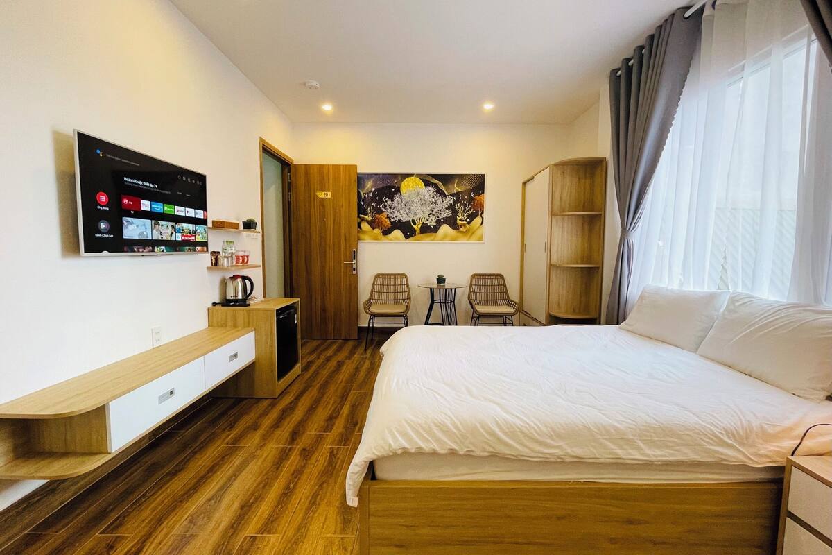 A hotel room with a bed, a TV mounted on the wall, a painting above the bed, a wooden floor, and a window with curtains.