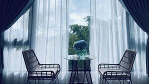 Una habitación con dos sillas de mimbre y una mesa de vidrio, ventanas amplias con cortinas translúcidas y vistas a zonas verdes.