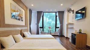 Habitación de hotel con dos camas, televisión, ventilador y vistas a árboles a través de amplias ventanas.