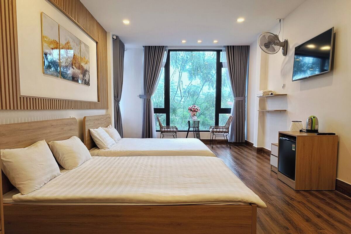 A hotel room with two beds, a TV, a fan, and a view of trees through large windows.