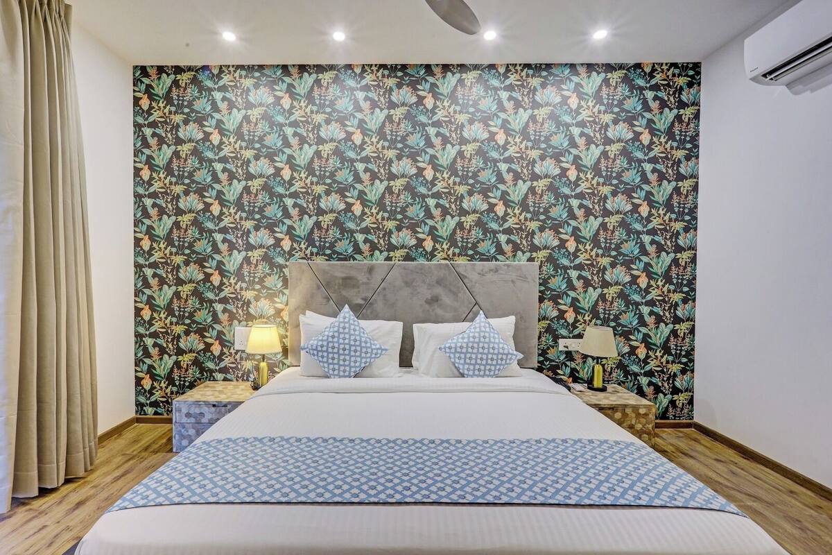 Un dormitorio con papel tapiz floral, una cama con almohadones estampados, mesitas de noche con lámparas y suelo de madera.