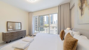A modern hotel room with a large bed, a dresser, a mirror, and a city view through large windows.