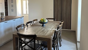 A dining area with a wooden table and chairs, a fruit bowl, and a kitchen in the background. - Charming air-conditioned house with garden and view on the Propriano bay (Olmeto)