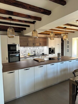 A modern kitchen with a large island, wooden beams on the ceiling, and pendant lighting. - Charming air-conditioned house with garden and view on the Propriano bay (Olmeto)