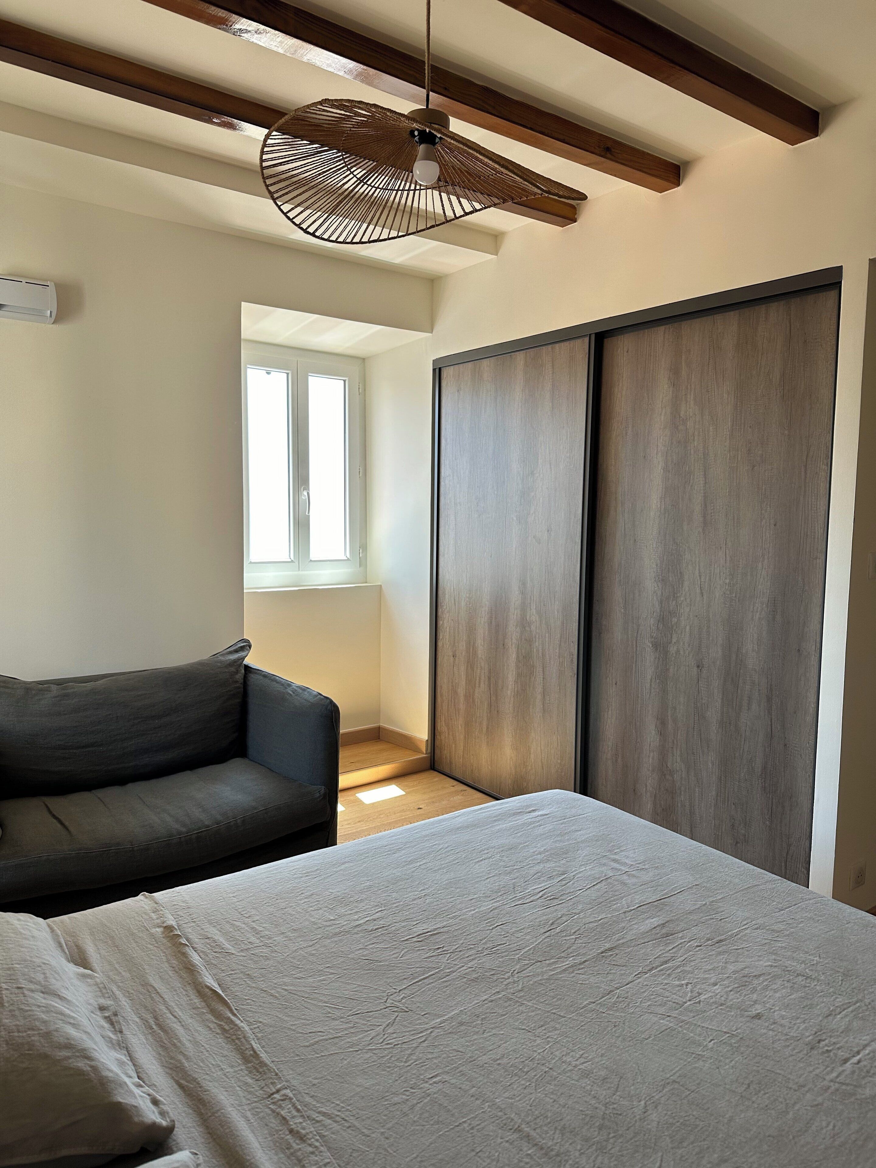A bedroom with a bed, a dark-colored sofa, a wooden wardrobe, and a hanging light fixture.
