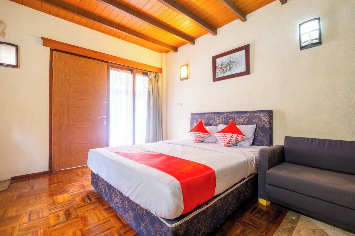 Un dormitorio con suelo de madera, una cama con manta roja, un sofá gris y techo de madera.