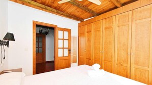 Une chambre avec un plafond en bois, un lit recouvert de linge de lit blanc, une armoire en bois et une porte donnant sur une autre pièce.