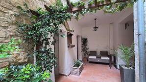 A cozy courtyard with a stone wall, a wooden pergola covered in greenery, a seating area with cushions, and a hanging lantern.