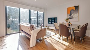 A modern living room with a sofa, dining table, and chairs, large windows, and a flat-screen TV. - StayAU Stylish 4BR Home Ringwood (Ringwood East)