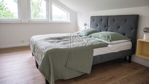 Une chambre à coucher avec un lit, une tête de lit rembourrée de couleur grise, une table de chevet avec une lampe et une fenêtre donnant sur un paysage verdoyant.