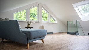 Un salon moderne avec un canapé gris, un plancher en bois et de grandes fenêtres donnant sur un paysage verdoyant.
