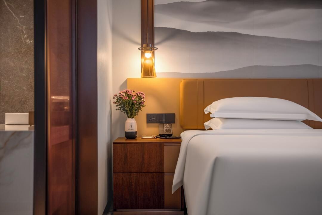 A hotel room with a bed, bedside table, lamp, and a painting of a mountain landscape.