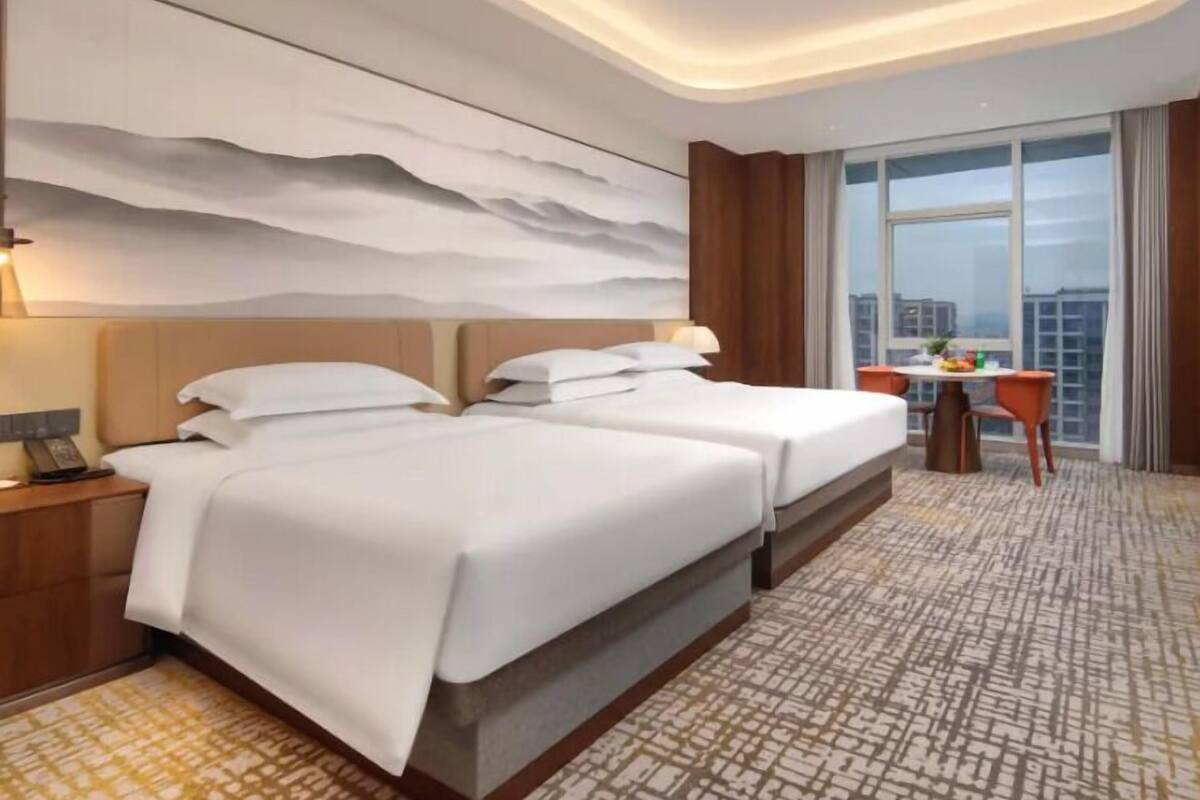 A hotel room with two beds, a large window with a view, a desk with a chair, and a patterned carpet.
