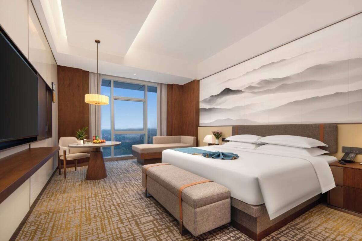 A modern hotel room with a large bed, a desk, a dining area, and a large window offering a city view.