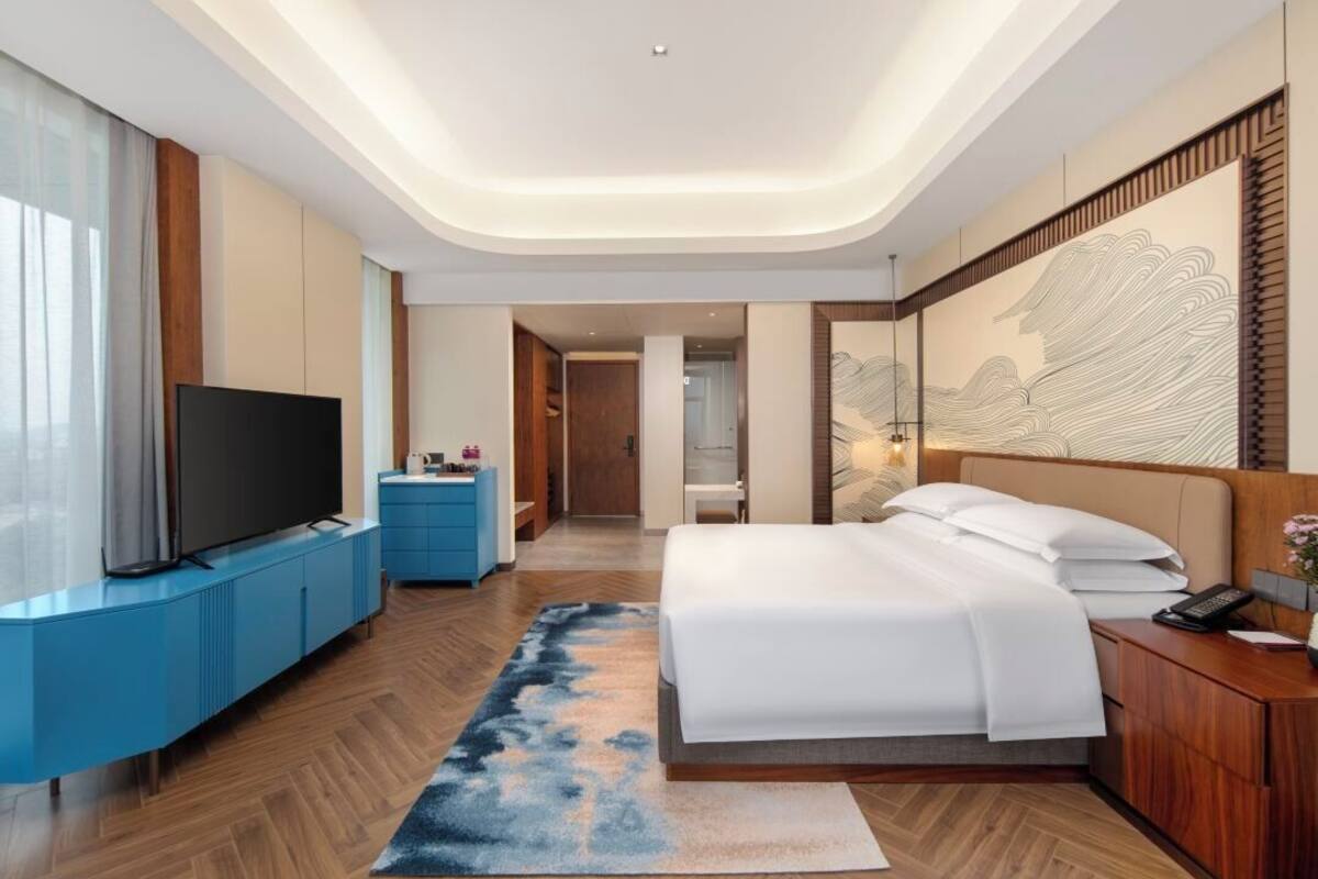 A modern hotel room with a flat-screen TV, a bed, a desk, and a large artwork on the wall.