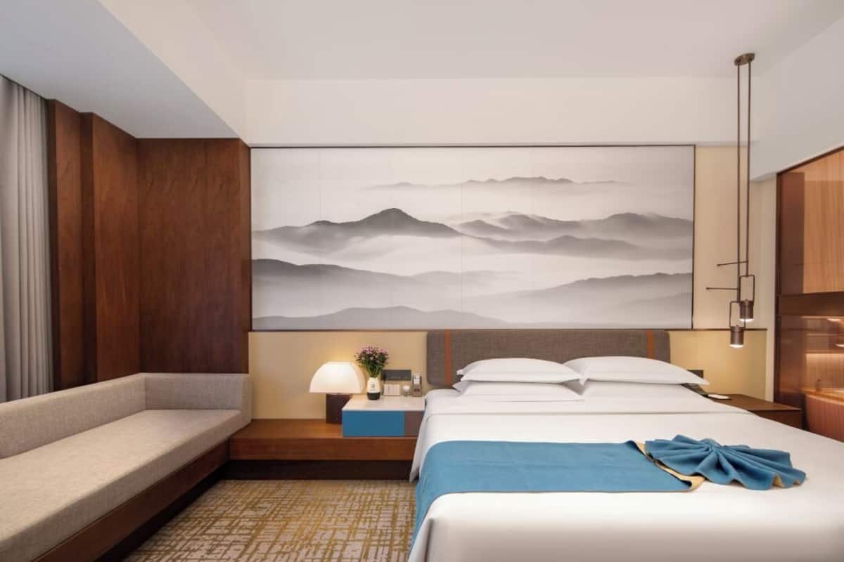 A hotel room with a large bed, a sofa, a nightstand, a lamp, and a wall-mounted artwork of mountains.