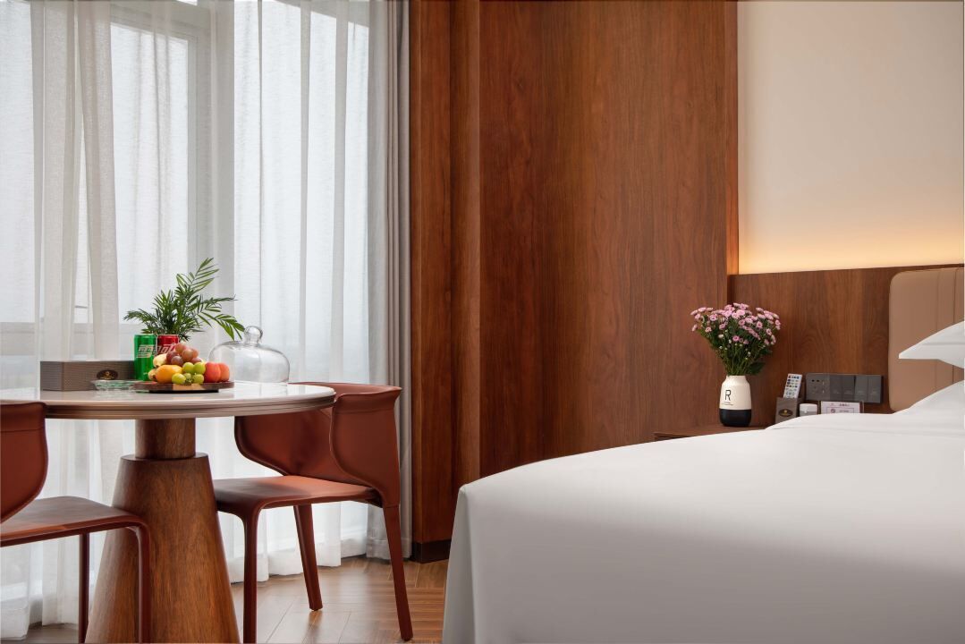 A hotel room with a bed, a dining table with chairs, a fruit bowl, a vase with flowers, and a wooden panel wall.