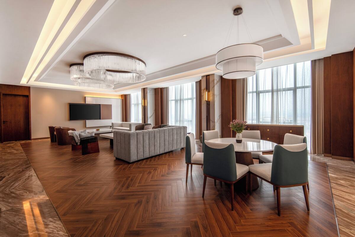 A spacious hotel lobby with a herringbone wood floor, a central seating area with a sofa and chairs, a dining table with chairs, and a large chandelier.