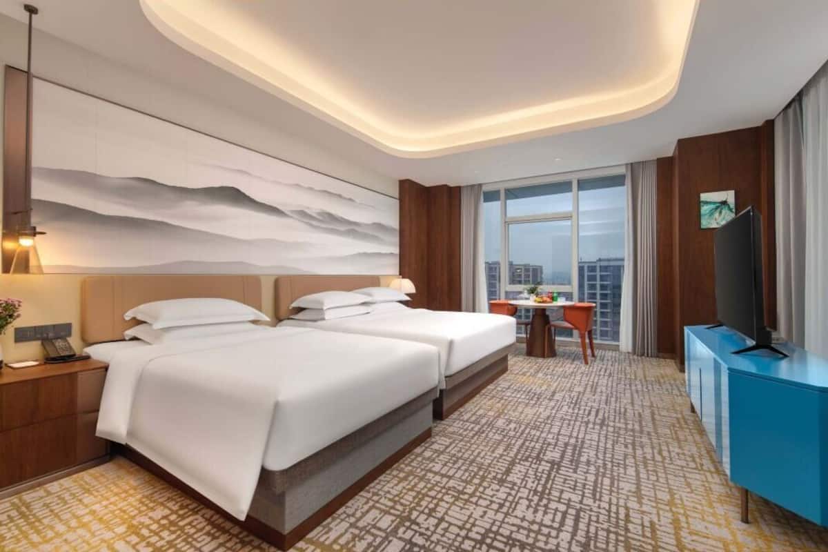 A hotel room with two beds, a desk, a TV, and a large window offering a city view.