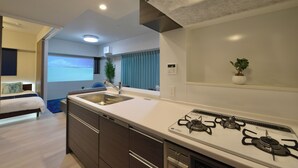 A modern kitchen with a gas stove, a sink, and a dishwasher. There is a bed with a blue headboard in the adjacent room. - Branchera Naha Akebono Premist 1206 (Naha)