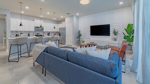 A modern living room with a sofa, a TV mounted on the wall, and a kitchen area with white cabinets and a microwave. - Stylish 5BR Retreat with Pool Near Disney (Davenport)