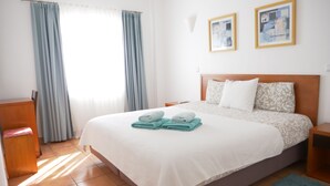 Une chambre d’hôtel comprenant un lit, une table de chevet, une chaise, une fenêtre avec des rideaux et deux tableaux encadrés accrochés au mur.