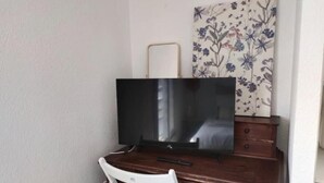 Ett litet, privat rum med ett skrivbord i trä, en platt-TV, en vit stol och ett blommönstrat konstverk på väggen.
