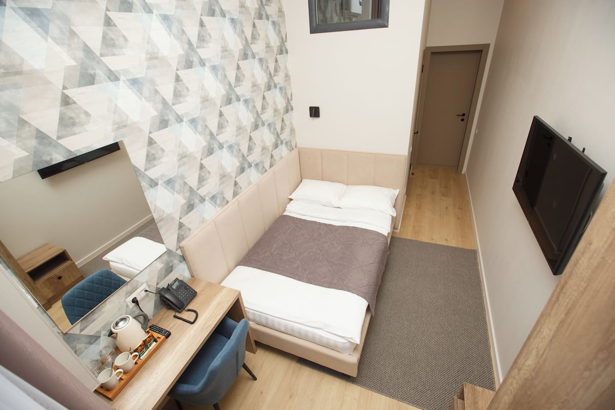 A compact hotel room with a bed, a desk with a chair, a small table with a tray of items, and a television mounted on the wall.