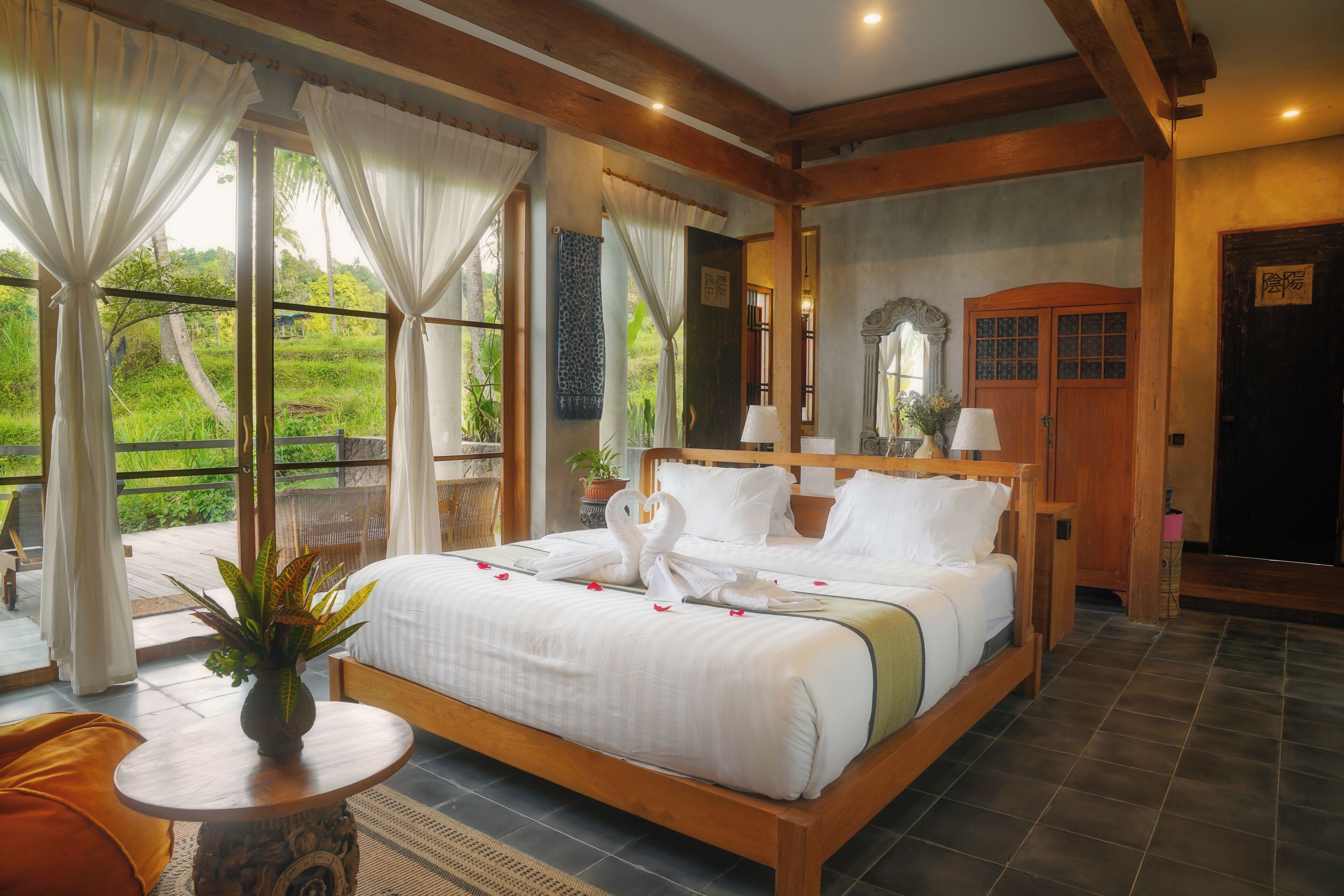 A spacious bedroom with a large bed, a small table with a plant, and a view of greenery outside.