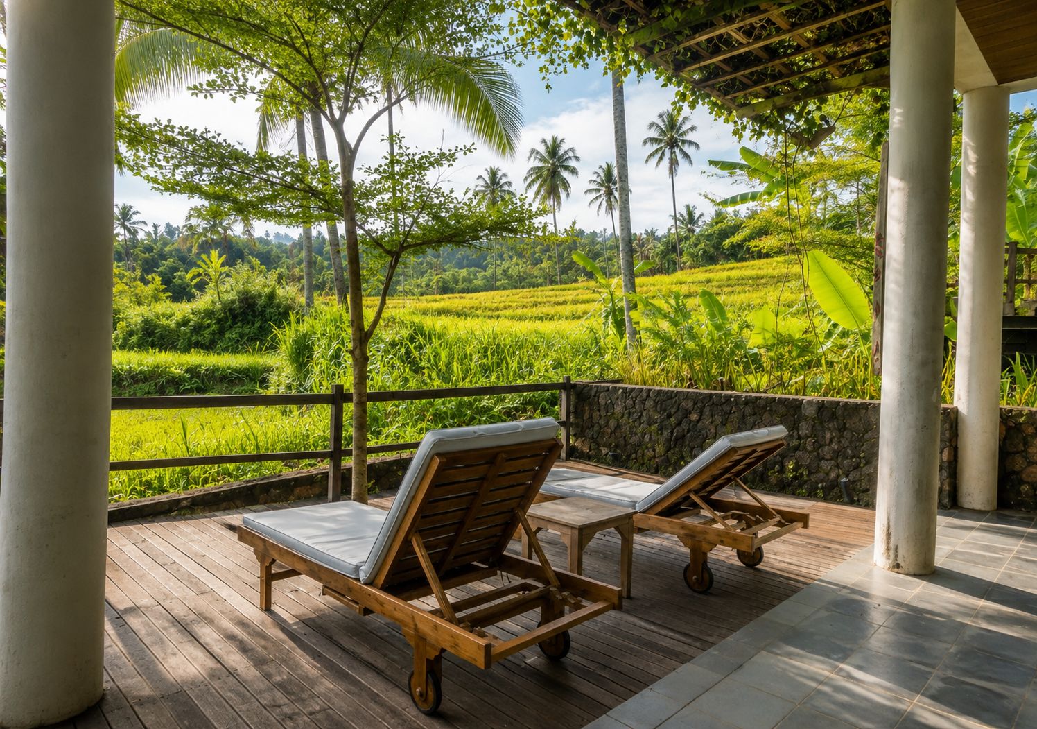 Una terraza de madera con dos sillones de descanso, un muro de piedra y vistas a un exuberante paisaje verde con palmeras.