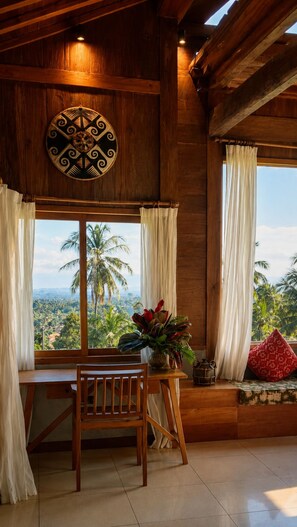 A wooden interior with a table and chairs, a decorative wall hanging, and a view of palm trees through a window. - Villa OMantra Home Bali (Sukasada)