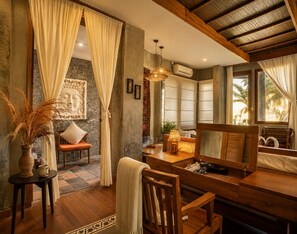 A room with a wooden desk, a chair, a sofa, a window with a view of palm trees, and a decorative wall piece. - Villa OMantra Home Bali (Sukasada)
