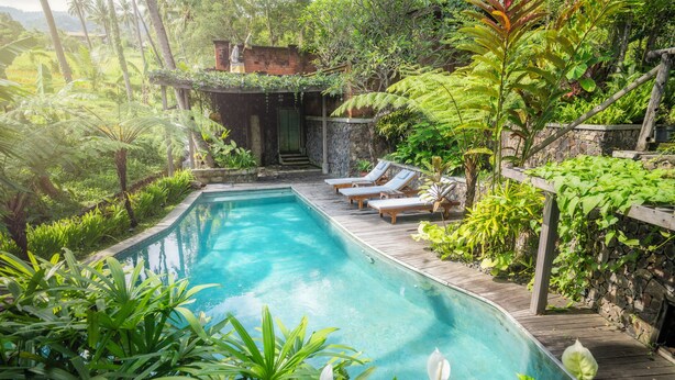 A swimming pool surrounded by lush greenery, wooden deck, and a stone wall. - Villa OMantra Home Bali (Sukasada)