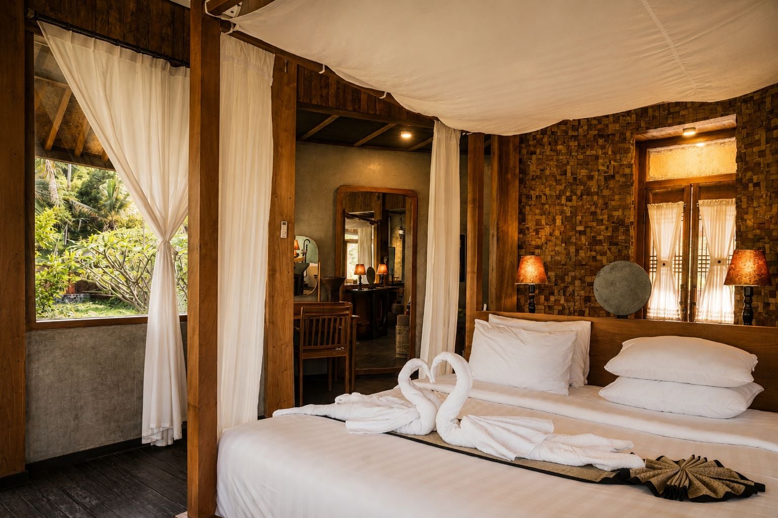 Una habitación con una cama, cortinas blancas, techo de madera, un espejo y vistas a la vegetación exterior.