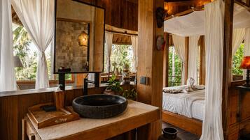 Una cabaña de madera con un espejo grande, un lavabo de piedra, una cama con dosel y vistas al verdor exterior.